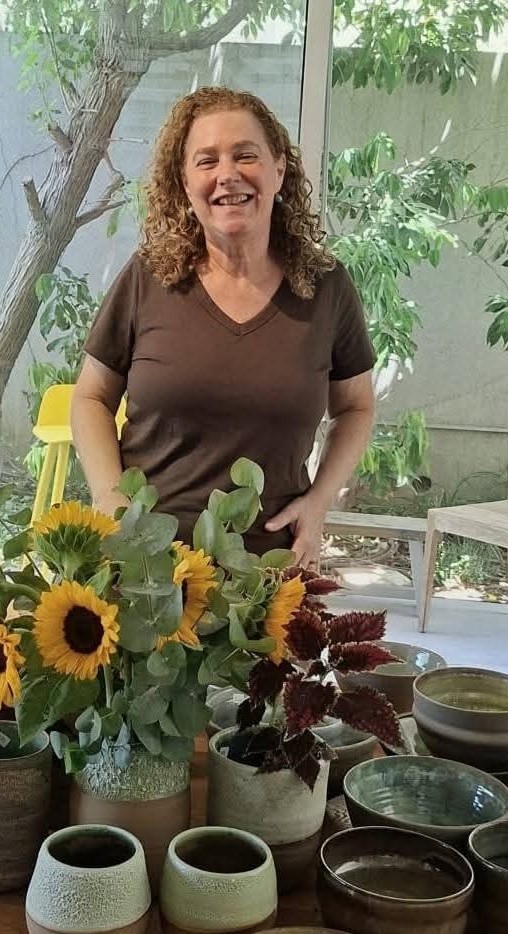 The maker of Marakima, smiling behind a table of her stoneware vessels and a vase of sunflowers.